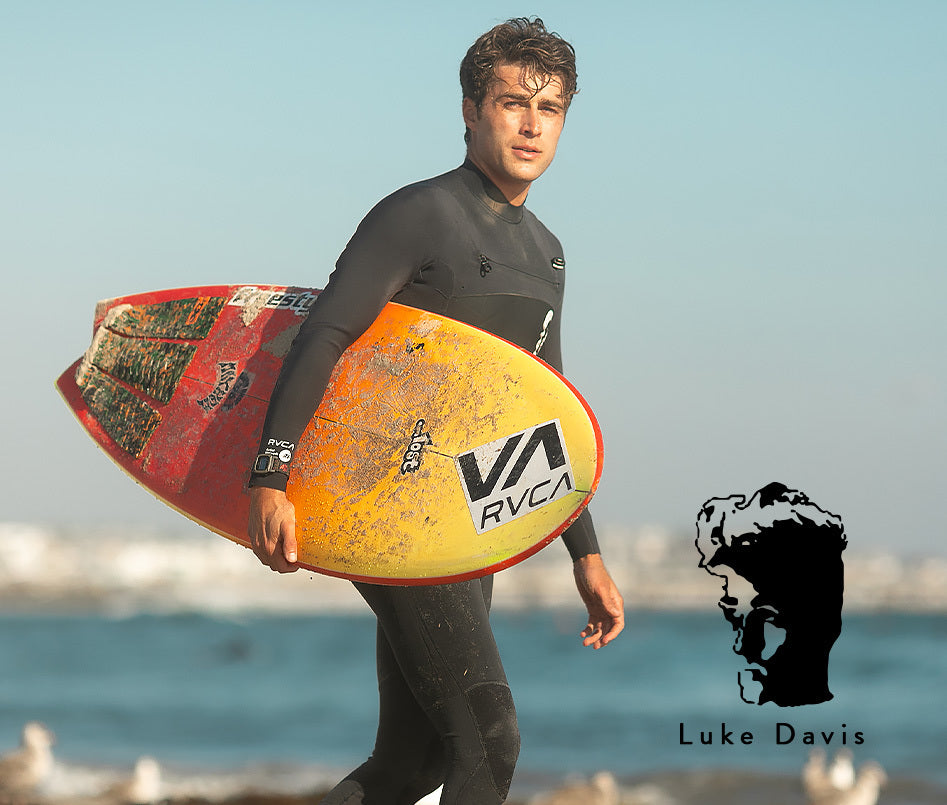 Luke Davis carrying a surfboard on the beach with a logo